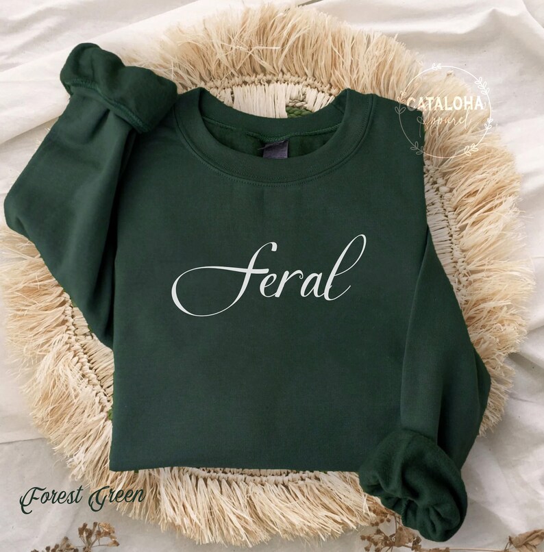 K&ouml;nnte beinhalten: Gr&uuml;n-farbener Sweatshirt mit dem Wort "feral" in wei&szlig;er, kursiv geschriebener Schrift.