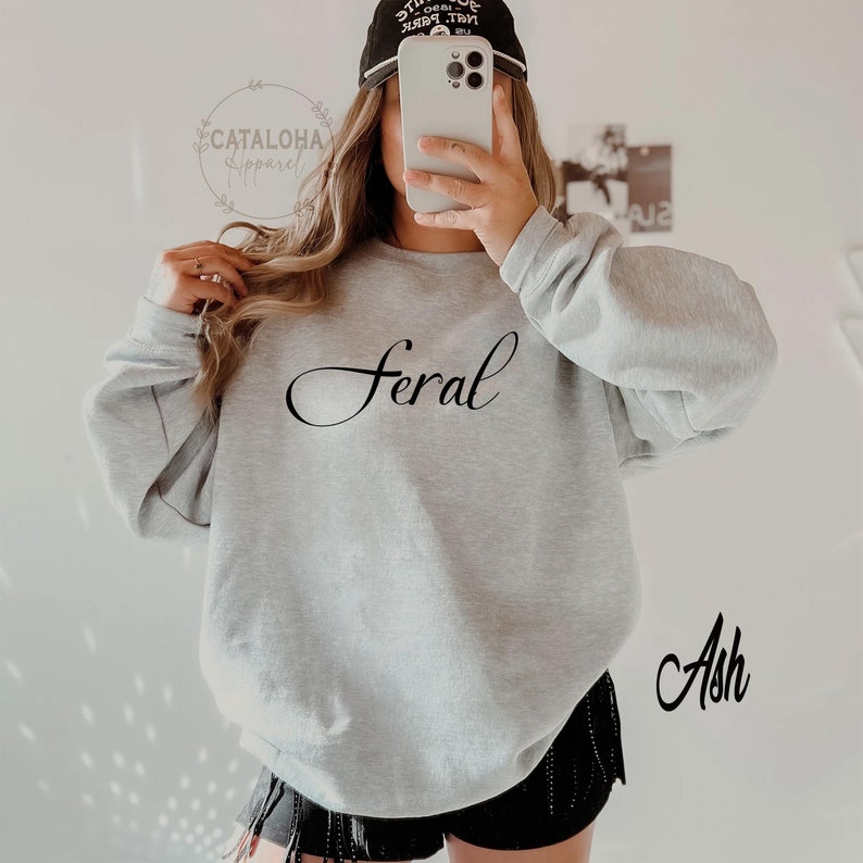 K&ouml;nnte beinhalten: Ein grauer Pullover mit dem Wort "Feral" in schwarzer Kursivschrift. Der Pullover wird von einer Person getragen und das Wort "Ash" ist in schwarzer Kursivschrift in der rechten unteren Ecke des Bildes geschrieben.