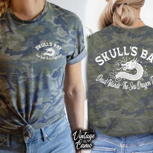 May include: A camouflage-patterned t-shirt with the text "Skull's Bay" and a dragon graphic. The shirt is olive green, blue, and gray. The back of the shirt also features the text "Skull's Bay" and a dragon graphic.