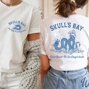 May include: White t-shirts with blue graphic designs. One shirt features "Skull's Bay" text with a dragon and the text "Dead Islands - The Sea Dragon Tavern". The other shirt has the same design on the back.