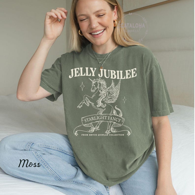 May include: An olive green t-shirt with the text "JELLY JUBILEE" above a winged unicorn graphic. Below the graphic is the text "STARLIGHT FANCY" on a banner. The shirt also has the word "Moss" printed on the bottom left.