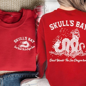 May include: Red sweatshirt with white graphic text and a dragon illustration. The text reads "SKULL'S BAY" and "Dead Islands - The Sea Dragon Tavern". The back features a ship, dragon, and text.
