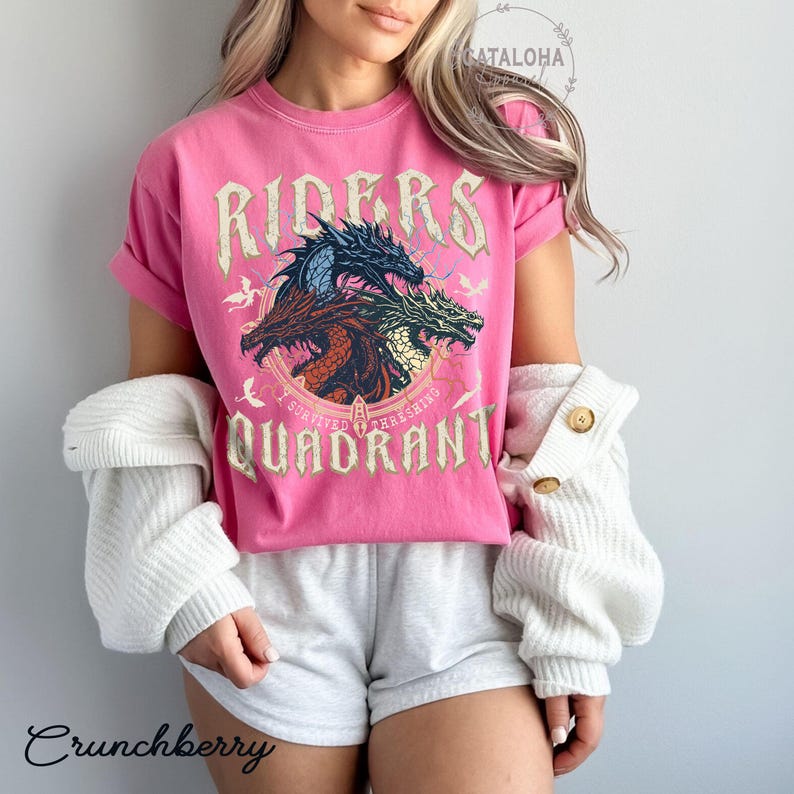May include: A pink t-shirt with a graphic of three dragons and the text "RIDERS QUADRANT". The shirt has rolled-up sleeves and a vintage style. The shirt is paired with gray shorts and a white cardigan.