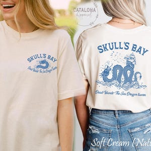 May include: Soft cream-colored t-shirt with a blue graphic on the front and back. The front graphic says "SKULL'S BAY" with a blue dragon. The back graphic features the same text and dragon design, with a ship and birds.