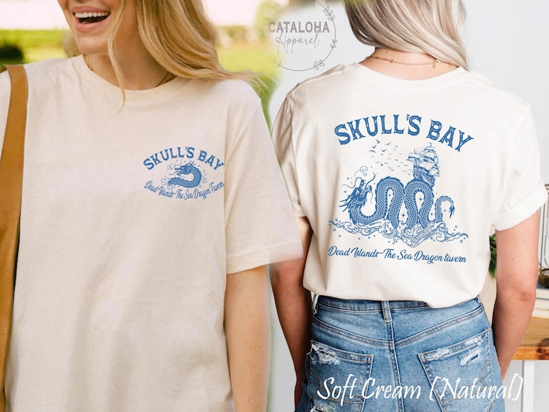 May include: Soft cream-colored t-shirt with a blue graphic on the front and back. The front graphic says "SKULL'S BAY" with a blue dragon. The back graphic features the same text and dragon design, with a ship and birds.