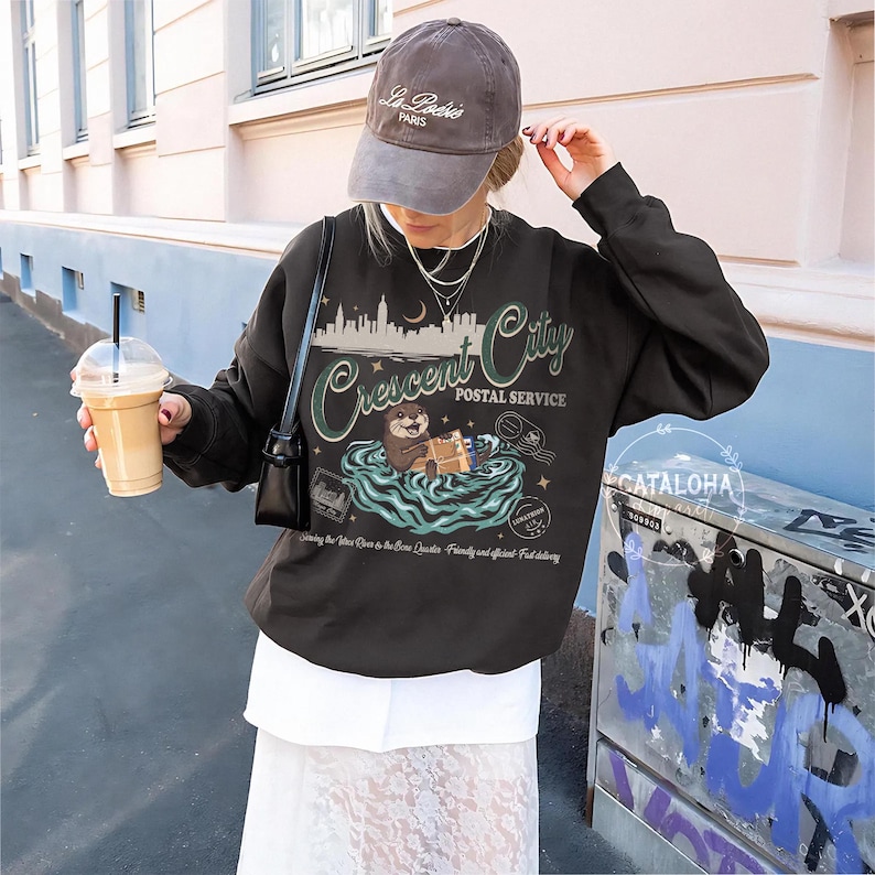 May include: A dark gray sweatshirt with a graphic featuring an otter delivering mail in a river, with the text "Crescent City Postal Service." The design includes a cityscape, moon, and the phrase "Serving the River to the Bone Quarter." A gray baseball cap is worn.