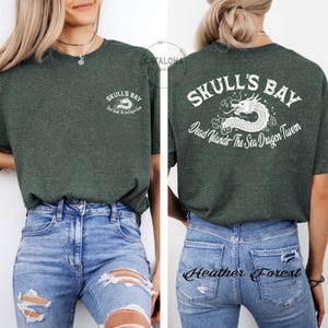 May include: Dark green t-shirt with the text "SKULL'S BAY" and a white dragon graphic on the front and back. The back of the shirt also includes the text "Dead Islands The Sea Dragon Tavern". The shirt is paired with blue jeans.