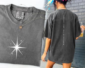 May include: A charcoal gray short-sleeve t-shirt with a vintage wash. The front features a white starburst graphic. The back displays vertical white text that reads "THROUGH THE DARK I WILL SHINE". The shirt has a crew neckline and a relaxed fit.