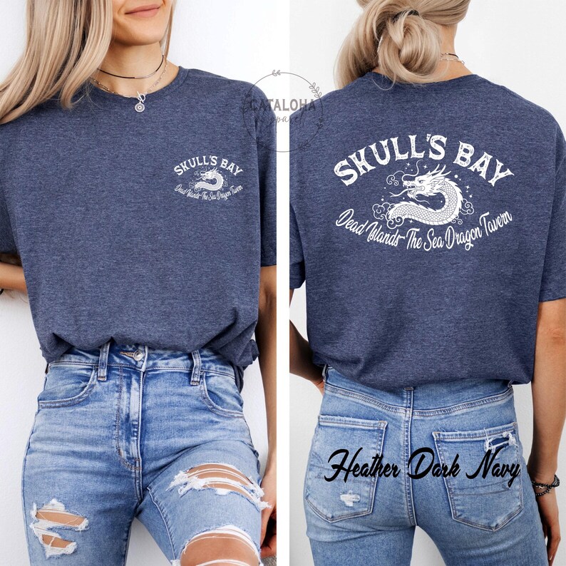 May include: Heather dark navy t-shirt with the text "SKULL'S BAY" and a white dragon graphic. The back of the shirt also features the text "Dead Islands - The Sea Dragon Tavern". The model is wearing ripped blue jeans.
