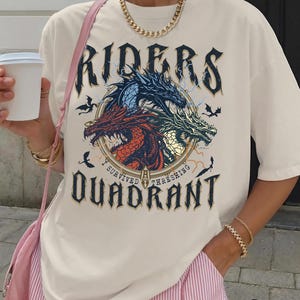 May include: Ivory-colored t-shirt featuring a graphic with three dragons in red, blue, and green, with the text "RIDERS" above and "I SURVIVED THRESHING QUADRANT" below. The shirt has short sleeves.