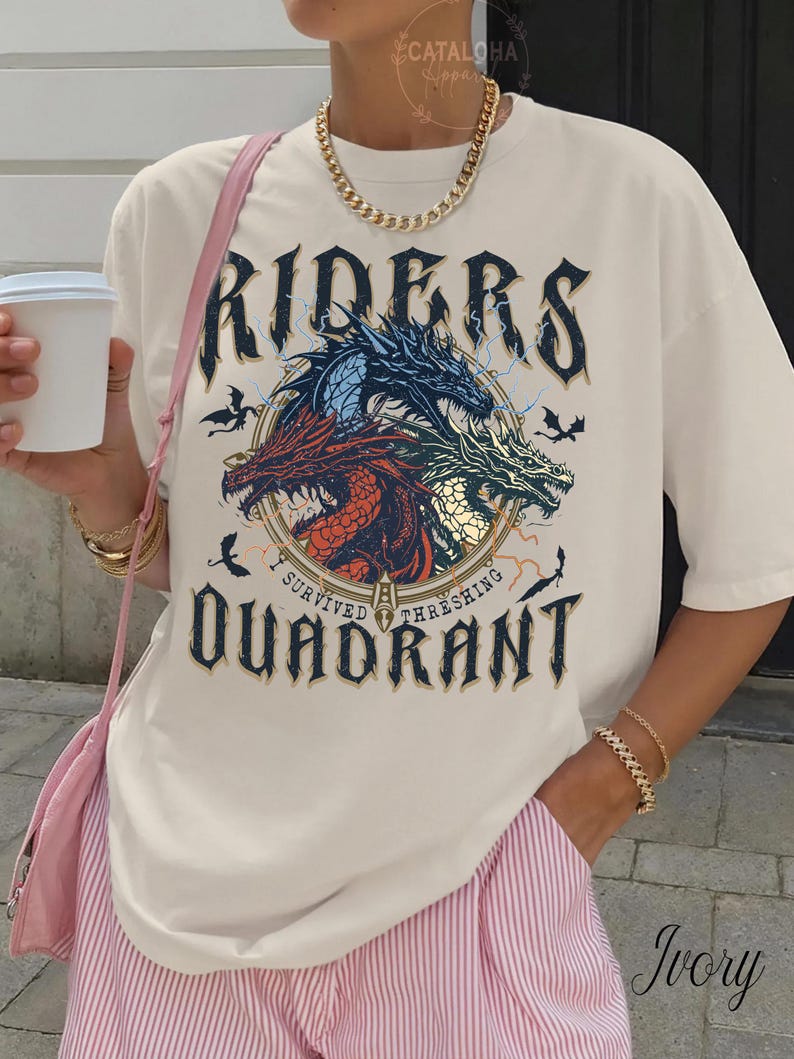 May include: Ivory-colored t-shirt featuring a graphic with three dragons in red, blue, and green, with the text "RIDERS" above and "I SURVIVED THRESHING QUADRANT" below. The shirt has short sleeves.