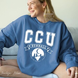 Pode incluir: Camisola azul &iacute;ndigo com letras brancas em bloco que dizem "CCU" acima de "SUNBALL" e um gr&aacute;fico de uma paisagem urbana com uma lua e um lobo. O texto "THE PACK OF DEVILS - LUNATHION" est&aacute; abaixo.
