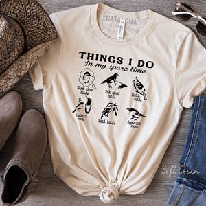 May include: A cream-colored t-shirt with the text "THINGS I DO In my spare time" and illustrations of birds. The shirt features various bird-related activities such as "Think about birds," "Talk about birds," and "Watch birds."