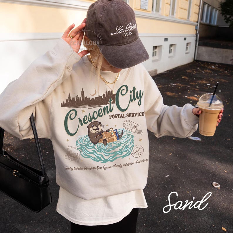 Op de afbeelding: Een zandkleurig sweatshirt met een afbeelding van een otter, de woorden "Crescent City Postal Service" en een stadsgezicht. Het sweatshirt wordt gecombineerd met een bruine baseballcap met de tekst "La Paris".