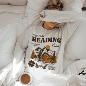 May include: A light grey sweatshirt featuring the words "Cozy Cabin Reading Club" and a graphic of a cabin, mountains, and trees. The sweatshirt is on a white bed with a cup of coffee.