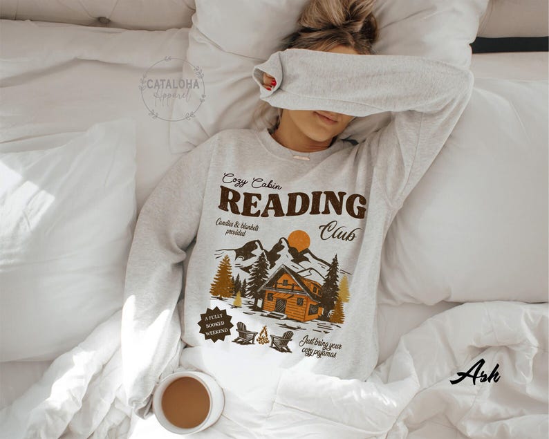 May include: A light grey sweatshirt featuring the words "Cozy Cabin Reading Club" and a graphic of a cabin, mountains, and trees. The sweatshirt is on a white bed with a cup of coffee.
