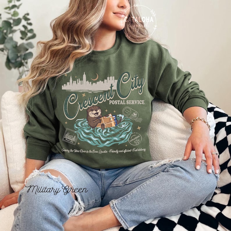 Op de afbeelding: Militairgroen sweatshirt met een afbeelding van een otter die post bezorgt in een rivier, met de tekst "Crescent City Postal Service". Het ontwerp bevat een stadsgezicht en de zin "Serving the Otter River to the Bone Quarter - Friendly and efficient - Fast delivery."
