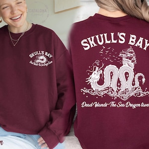 May include: Maroon sweatshirt with white graphic text and design. The front features "Skull's Bay" and a sea dragon. The back displays "Skull's Bay" with a ship and dragon illustration. Text reads "Dead Islands- The Sea Dragon Tavern."