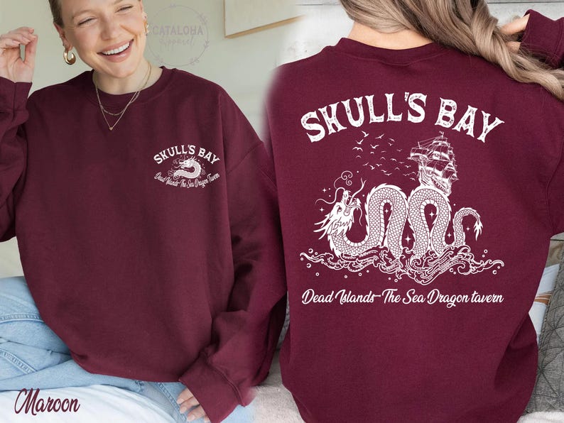 May include: Maroon sweatshirt with white graphic text and design. The front features "Skull's Bay" and a sea dragon. The back displays "Skull's Bay" with a ship and dragon illustration. Text reads "Dead Islands- The Sea Dragon Tavern."
