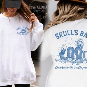 May include: White sweatshirt with blue graphic text and design. The front features "Skull's Bay" and "Dead Islands - The Sea Dragon Tavern". The back has a detailed illustration of a ship, dragon, and waves, with the same text.