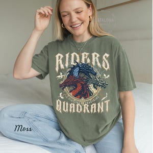 May include: Moss green t-shirt featuring a graphic of three dragons in red, blue, and white, with the words "RIDERS QUADRANT" in a vintage font. The shirt has a relaxed fit and short sleeves.