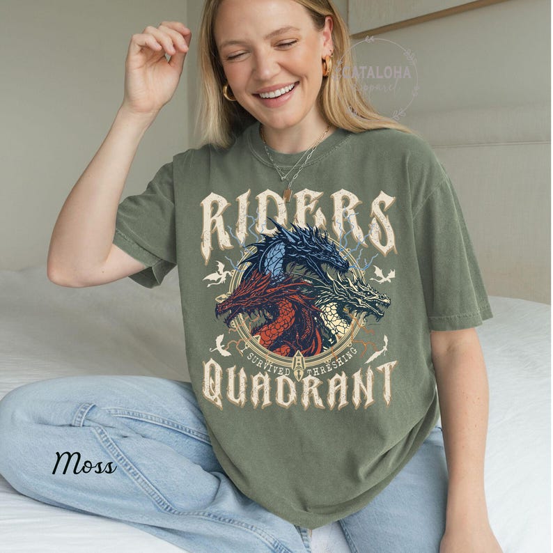 May include: Moss green t-shirt featuring a graphic of three dragons in red, blue, and white, with the words "RIDERS QUADRANT" in a vintage font. The shirt has a relaxed fit and short sleeves.