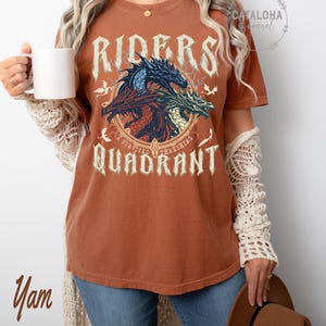 May include: A rust-colored t-shirt featuring a graphic of three dragons in blue, red, and green, with the words "RIDERS" and "QUADRANT" in a distressed font. The text "I SURVIVED THRASHING" is also present. The shirt is worn by a person.