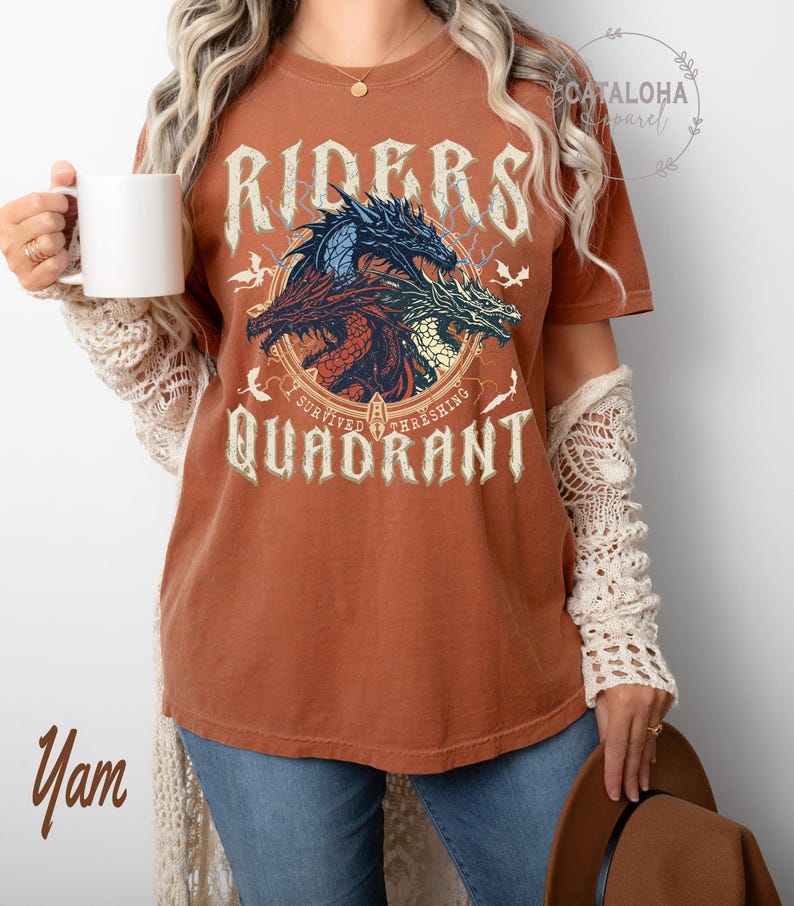 May include: A rust-colored t-shirt featuring a graphic of three dragons in blue, red, and green, with the words "RIDERS" and "QUADRANT" in a distressed font. The text "I SURVIVED THRASHING" is also present. The shirt is worn by a person.