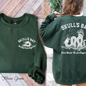 May include: Forest green sweatshirt with the text "SKULL'S BAY" and a white dragon graphic on the front and back. The back design includes a ship and the text "Dead Wands The Sea Dragon tavern".