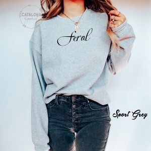K&ouml;nnte beinhalten: Ein graues Sweatshirt mit dem Wort "feral" in schwarzer Kursivschrift. Das Sweatshirt wird von einer Person getragen und der Text "Sport Grey" ist in der rechten unteren Ecke des Bildes sichtbar.
