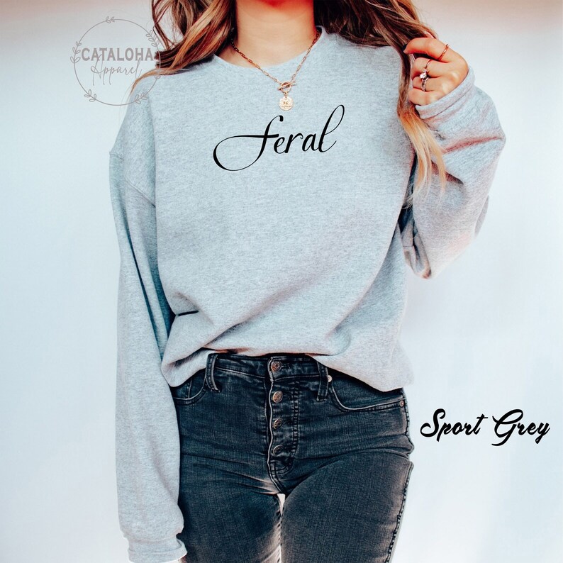 K&ouml;nnte beinhalten: Ein graues Sweatshirt mit dem Wort "feral" in schwarzer Kursivschrift. Das Sweatshirt wird von einer Person getragen und der Text "Sport Grey" ist in der rechten unteren Ecke des Bildes sichtbar.