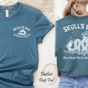 May include: Teal t-shirt with "SKULL'S BAY" text and a dragon graphic. The front of the shirt has the text "SKULL'S BAY" and "Dead Islands The Sea Dragon tavern". The back of the shirt has a dragon and ship graphic.