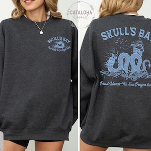 May include: Dark grey heather sweatshirt with "Skull's Bay" text and a sea dragon graphic in light blue on the front and back. The back design includes a ship and the text "Dead Islands - The Sea Dragon Tavern".