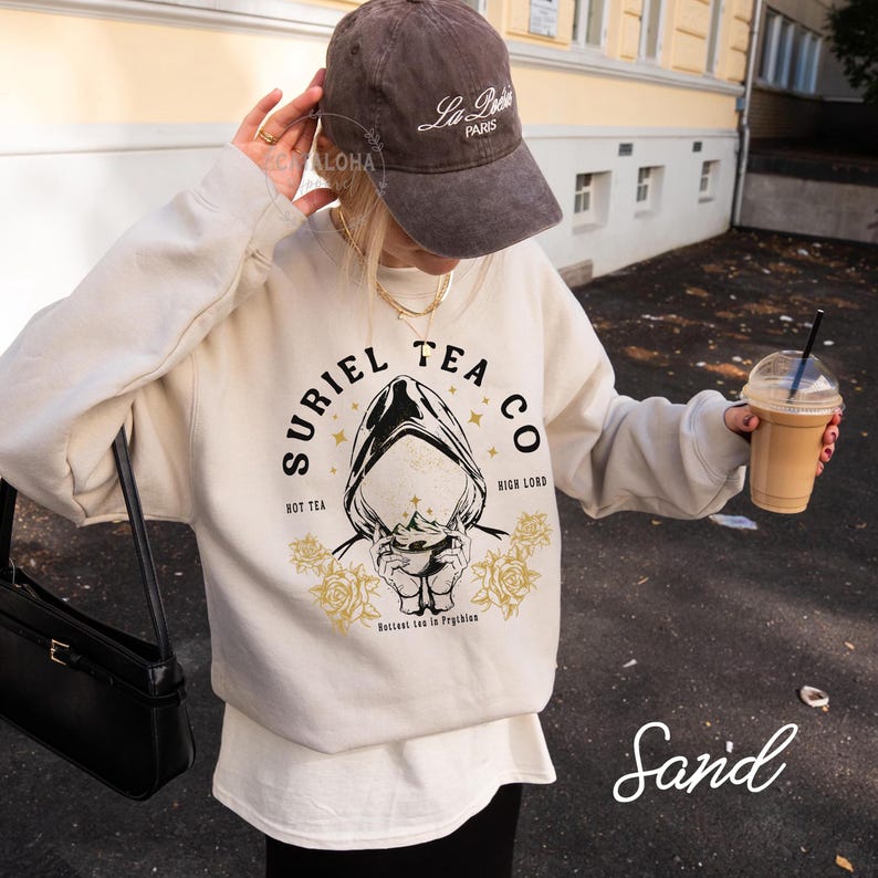 May include: Sand-colored sweatshirt with the text "SURIEL TEA CO" and a graphic of a hooded figure. The sweatshirt has a crew neck and long sleeves. A brown baseball cap with "La Paris" is worn. A black purse is visible.