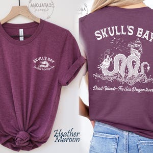 May include: Heather maroon t-shirt with "Skull's Bay" text and a dragon graphic on the front and back. The back design includes a ship and the text "Dead Islands The Sea Dragon tavern".