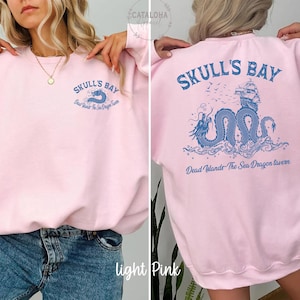 May include: Light pink sweatshirt with blue graphic text and design. The front features "SKULL'S BAY" and "Dead Islands The Sea Dragon Tavern". The back displays a blue dragon, ship, and text "SKULL'S BAY" and "Dead Islands The Sea Dragon Tavern".