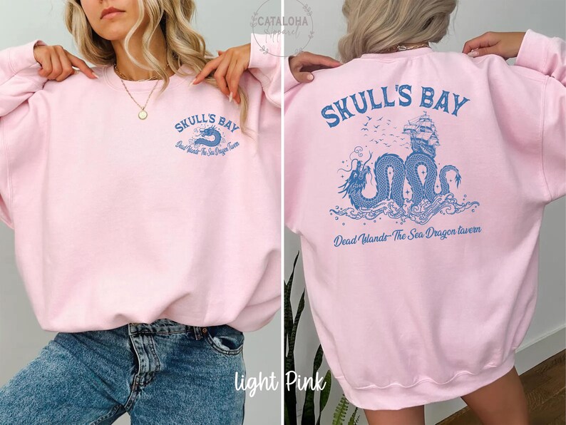 May include: Light pink sweatshirt with blue graphic text and design. The front features "SKULL'S BAY" and "Dead Islands The Sea Dragon Tavern". The back displays a blue dragon, ship, and text "SKULL'S BAY" and "Dead Islands The Sea Dragon Tavern".