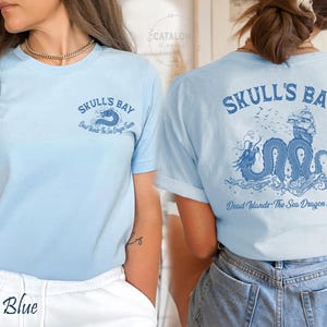 May include: Baby blue t-shirt with the text "SKULL'S BAY" and a dragon graphic on the front and back. The back graphic includes a ship and the text "Dead Islands - The Sea Dragon Tavern".