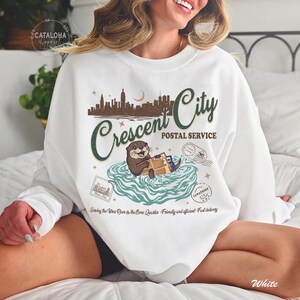 Op de afbeelding: Witte sweatshirt met een afbeelding van een otter die pakketten bezorgt, met de tekst "Crescent City Postal Service". Het ontwerp bevat een stadsgezicht, sterren en postzegels. De tekst "Sewing the Vitres River & the Bore Quarter - Friendly and efficient - Fast delivery" is ook opgenomen.