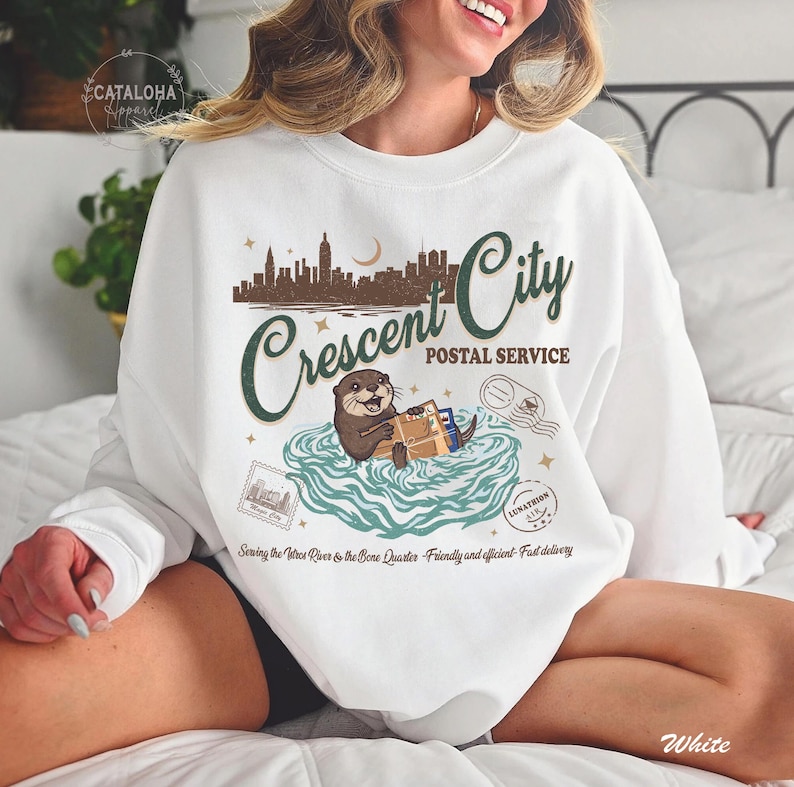 Op de afbeelding: Witte sweatshirt met een afbeelding van een otter die pakketten bezorgt, met de tekst "Crescent City Postal Service". Het ontwerp bevat een stadsgezicht, sterren en postzegels. De tekst "Sewing the Vitres River & the Bore Quarter - Friendly and efficient - Fast delivery" is ook opgenomen.