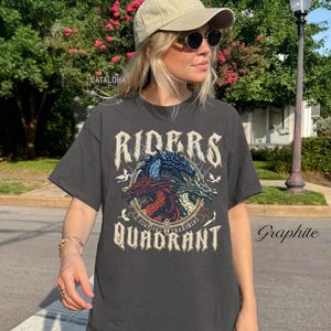 May include: Graphite-colored t-shirt featuring a dragon design with the words "Riders Quadrant". The graphic includes three dragons in red, blue, and green. The shirt is worn by a person wearing a cap and sunglasses.
