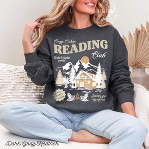 May include: Dark grey heather sweatshirt with a graphic that reads "Cozy Cabin Reading Club." The design features a cabin, mountains, trees, and the text "Candles & blankets provided." The bottom of the sweatshirt reads "A fully booked weekend" and "Just bring your cozy pyjamas."