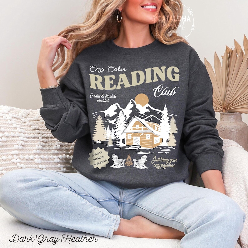 May include: Dark grey heather sweatshirt with a graphic that reads "Cozy Cabin Reading Club." The design features a cabin, mountains, trees, and the text "Candles & blankets provided." The bottom of the sweatshirt reads "A fully booked weekend" and "Just bring your cozy pyjamas."