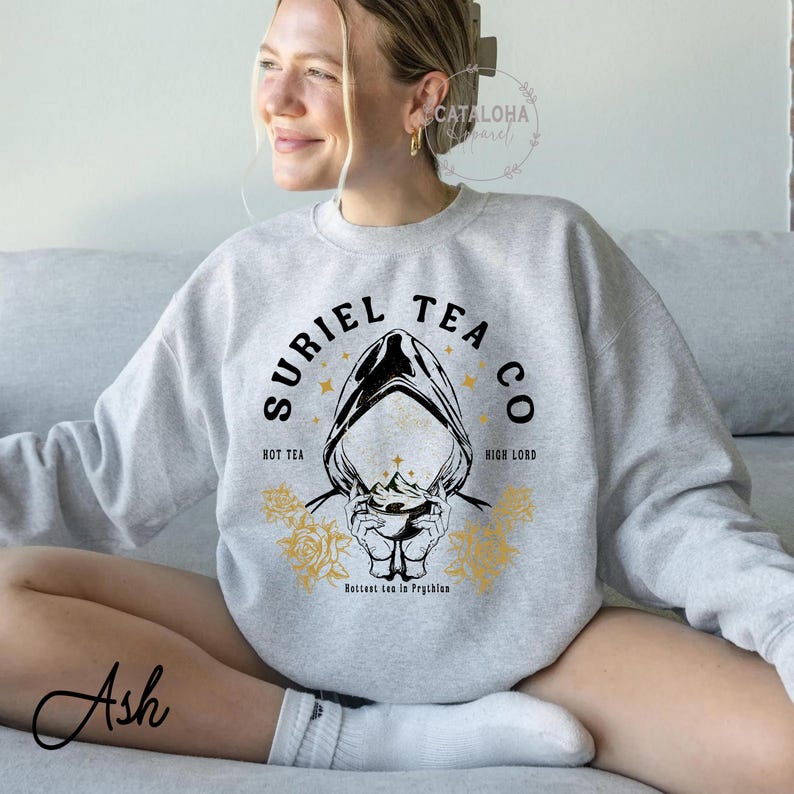 May include: A light gray sweatshirt with the text "SURIEL TEA CO" in a black, stylized font. The design includes a hooded figure, stars, and floral accents in gold. The words "Hot Tea" and "High Lord" are also present.