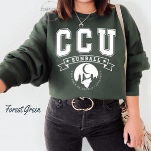 Pode incluir: Camisola verde floresta com letras brancas "CCU" acima de "SUNBALL". Abaixo, um gr&aacute;fico de uma paisagem urbana com um lobo uivando e o texto "THE PACK OF DEVILS - LUNATHION".