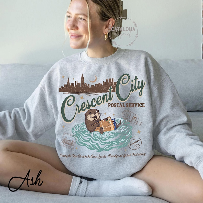 Op de afbeelding: Grijze sweatshirt met een afbeelding van een otter die post bezorgt, met de tekst "Crescent City Postal Service". Het ontwerp bevat een stadsgezicht, een maan en de zin "Serving the Vetra River & the Bene Quarter - Friendly and efficient - Fast delivery."