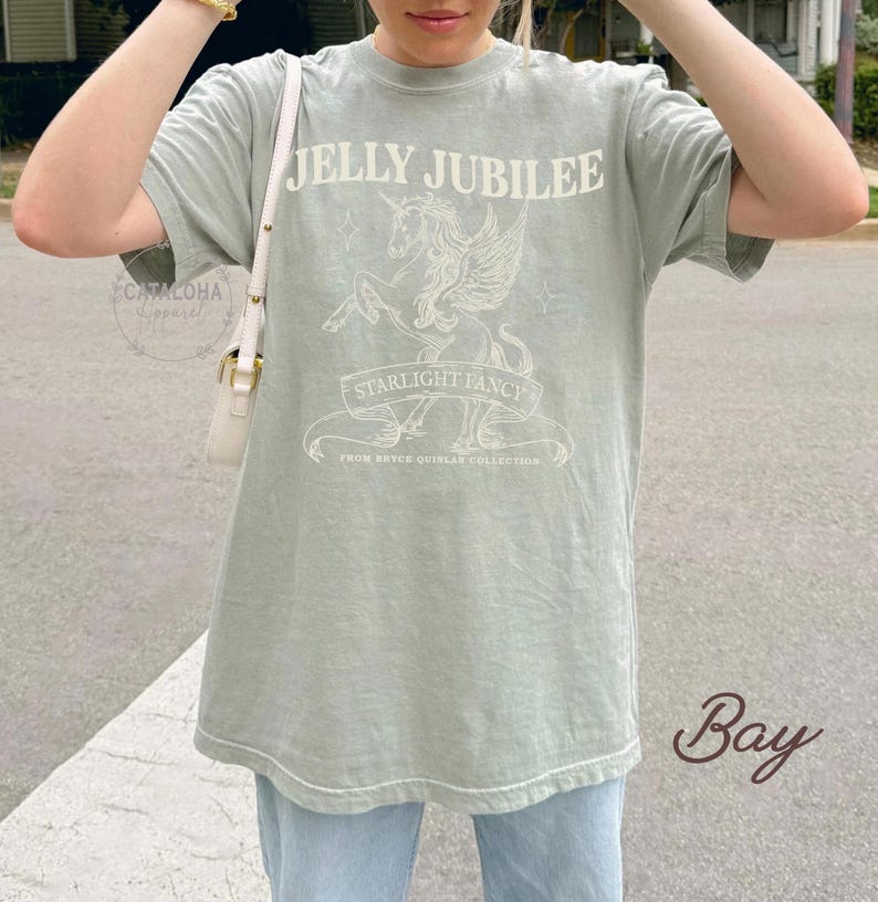 May include: Sage green t-shirt with the text "JELLY JUBILEE" above a unicorn graphic and the words "STARLIGHT FANCY" on a banner. The shirt also includes the text "FROM BRYCE QUINLAR COLLECTION".