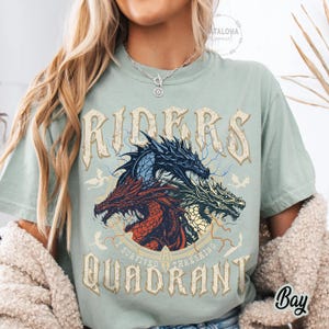 May include: A light green t-shirt featuring a graphic of three dragons in red, blue, and green, with the text "RIDERS QUADRANT" in a vintage font. The shirt also includes the text "I SURVIVED THRESHING".