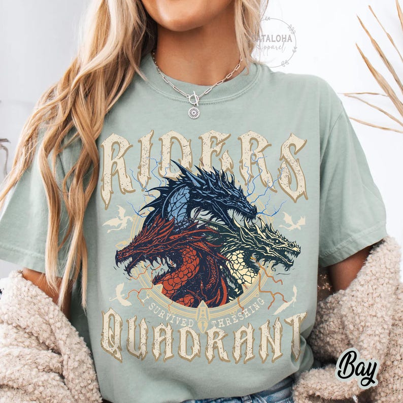 May include: A light green t-shirt featuring a graphic of three dragons in red, blue, and green, with the text "RIDERS QUADRANT" in a vintage font. The shirt also includes the text "I SURVIVED THRESHING".
