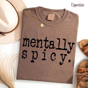 May include: Espresso-colored t-shirt with the phrase "mentally spicy" printed in black. A gold necklace with a pendant rests on the shirt. A cream-colored hat and sandals are also visible.
